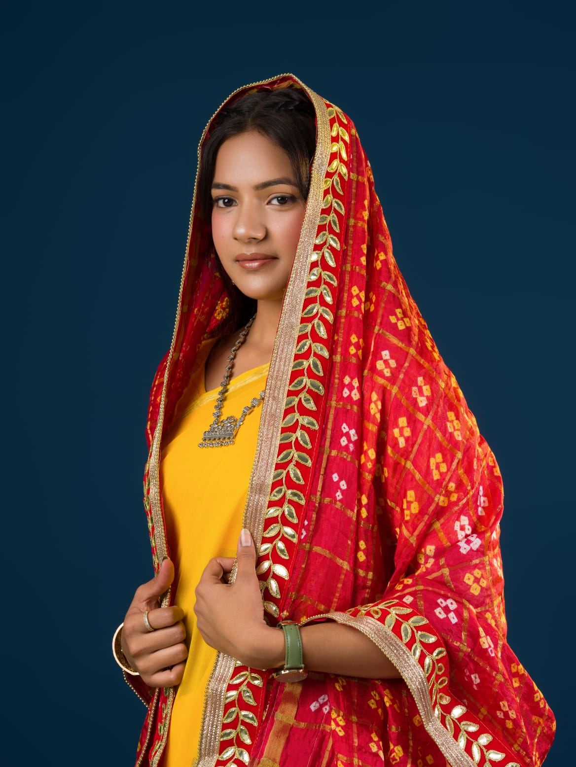 A woman in a traditional yellow attire is holding a red and yellow Bandhani silk chunri with floral patterns and a border.