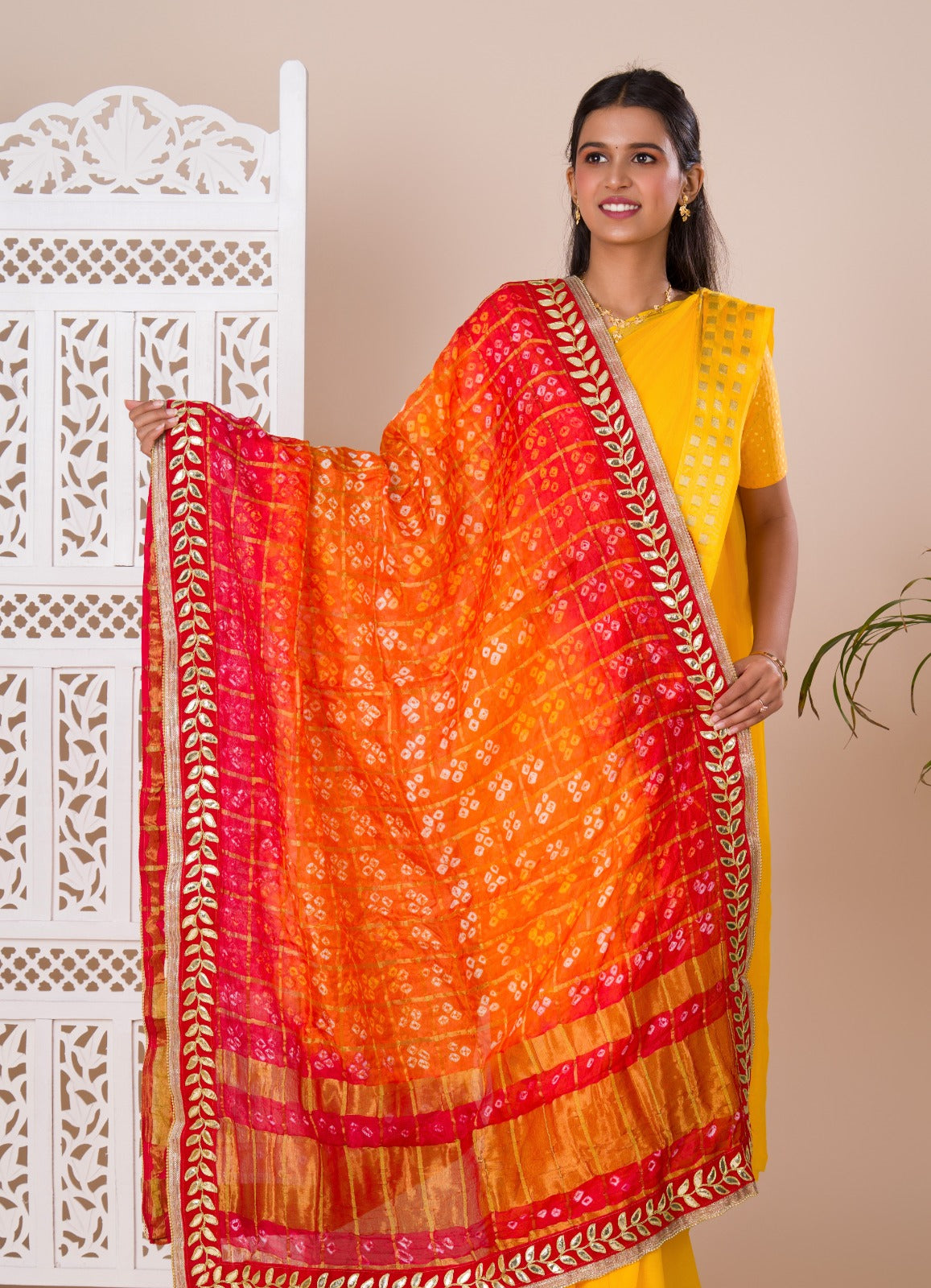 Atraditional Rajasthani Bandhani Chunri dupatta in silk, primarily in red and orange colors with a border, draped over her shoulders.