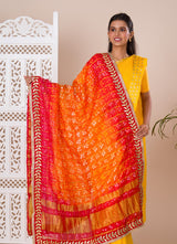 Atraditional Rajasthani Bandhani Chunri dupatta in silk, primarily in red and orange colors with a border, draped over her shoulders.