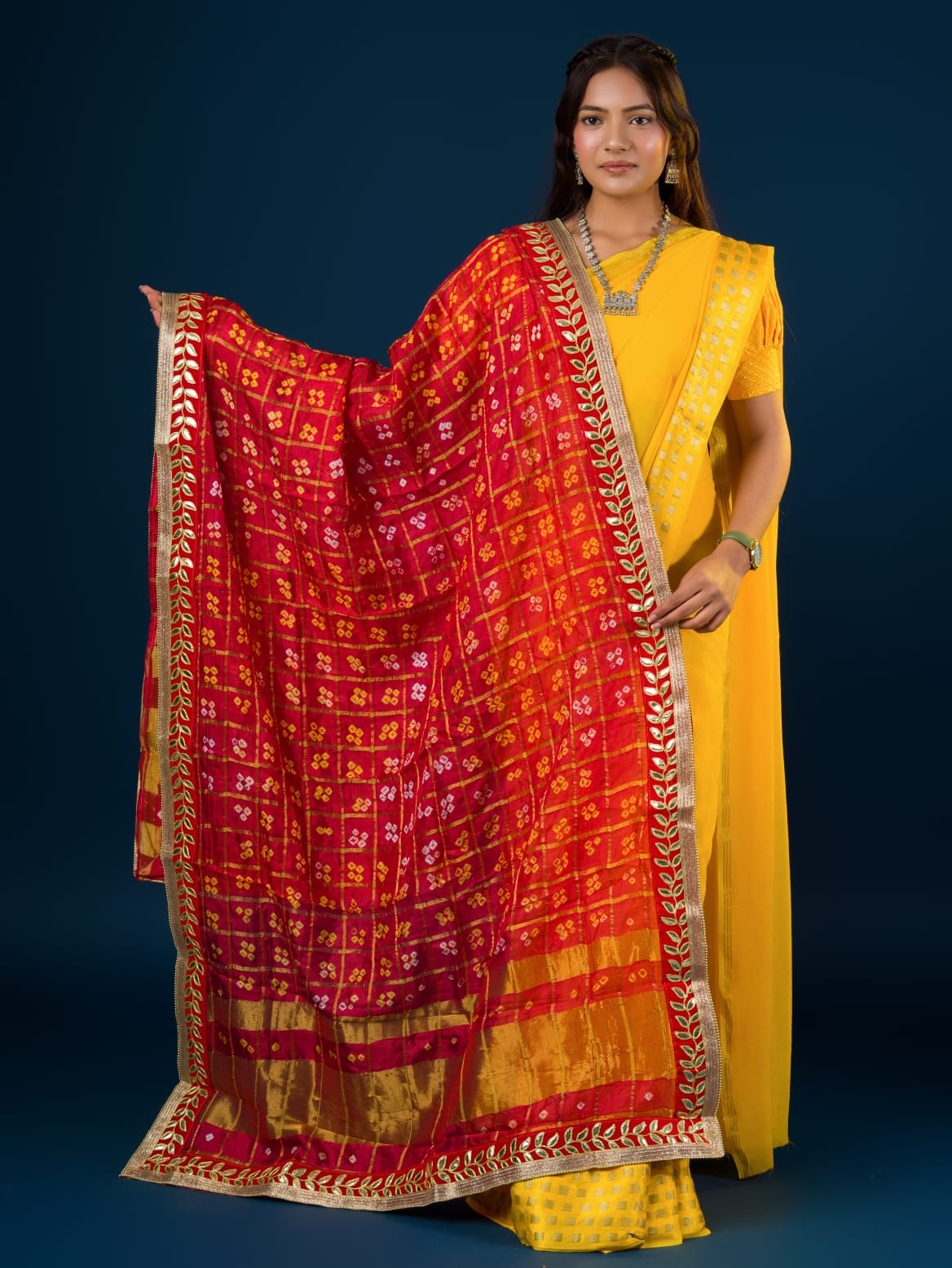A woman in a traditional yellow attire is holding a red and yellow Bandhani silk chunri with floral patterns and a border.