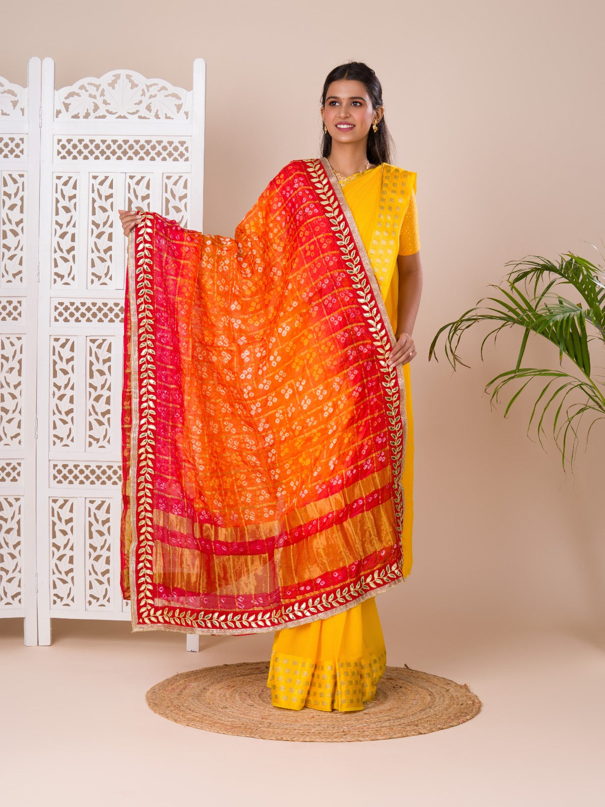 Atraditional Rajasthani Bandhani Chunri dupatta in silk, primarily in red and orange colors with a border, draped over her shoulders.